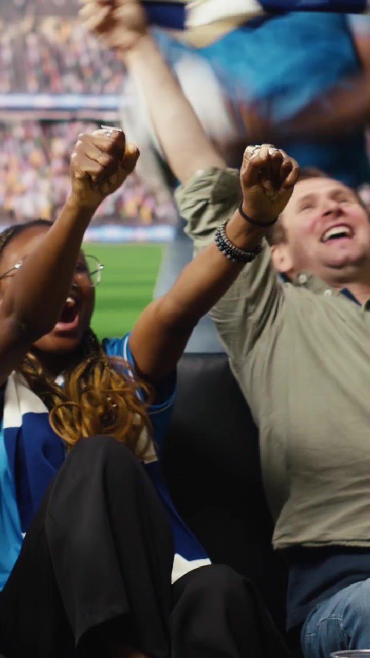 Video vertical: Aficionados al fútbol alegres en el sofá celebran un gol, sintiéndose como si estuvieran dentro del estadio