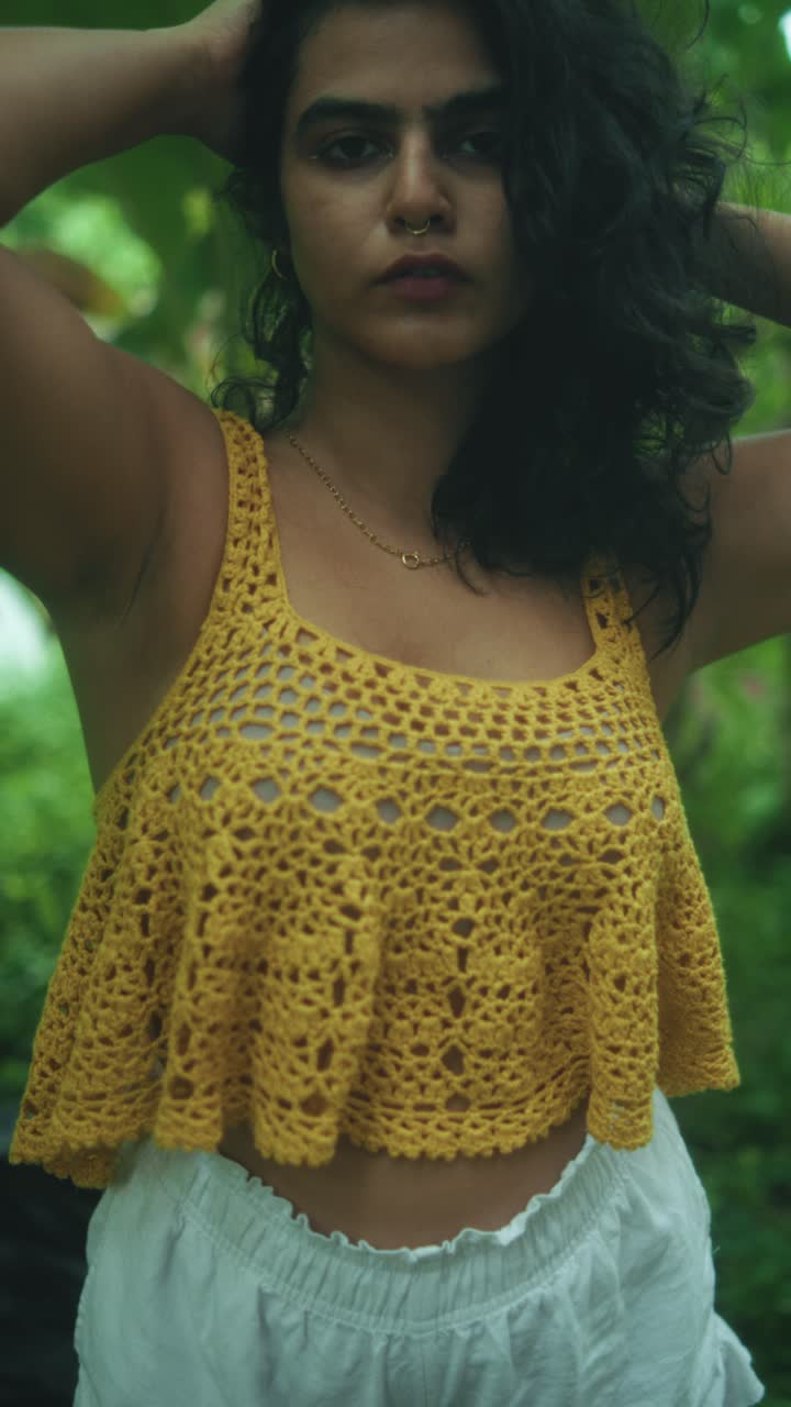 A woman wearing a yellow top and white shorts looks straight into the camera while posing confidently in an outdoor setting