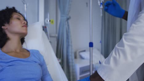 Diverse-male-doctor-preparing-drip-bag-for-female-patient-in-hospital-room