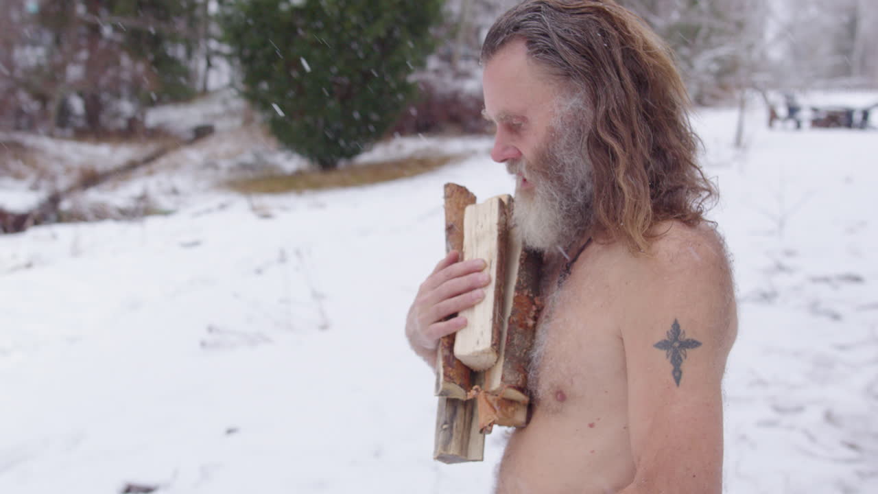 Scruffy Nordic man walks bare chested in cold snowy outdoors carrying firewood