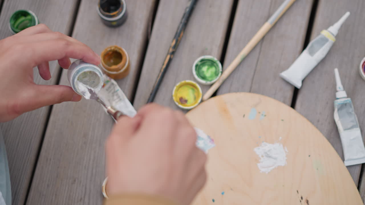 Kunstenaarshanden bereiden verf op een palet voor, potten met metallic groen en goud, penseel dat wit pigment laadt, houten promenade, ontspannen schetssessie in de buitenlucht met een gefocuste creatieve sfeer