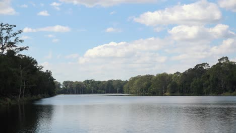 wide exterior time lapse shot of white clouds drifting above a breath taking scene of lake casting shadows over trees as they move in the blue sky in he day-time