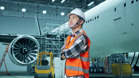 vista lateral de un ingeniero masculino asiático con casco de seguridad de pie con aviones en el hangar. cruzando los brazos mientras el mantenimiento de aviones