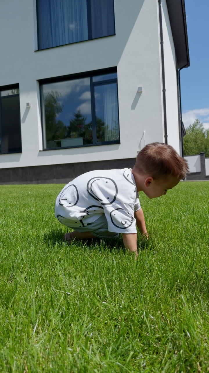 Lovely Caucasian baby boy sits squatted outdoors in the sun. Kid touches green soft grass with his hands. Vertical video.