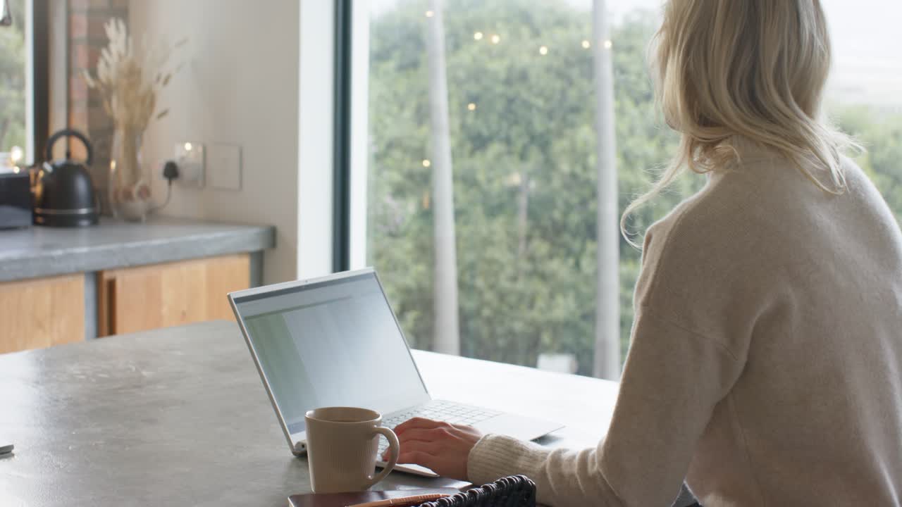 Placing hands on laptop typing at counter by window beside coffee mug, pausing to call smartphone
