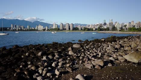 Flug-über-Die-Felsige-Küste-Am-Kitsilano-Beach-Mit-Ruhigem-Blauen-Meer,-Der-Skyline-Der-Innenstadt-Und-Dem-English-Bay-Beach-In-Vancouver,-Britisch-Kolumbien,-Kanada