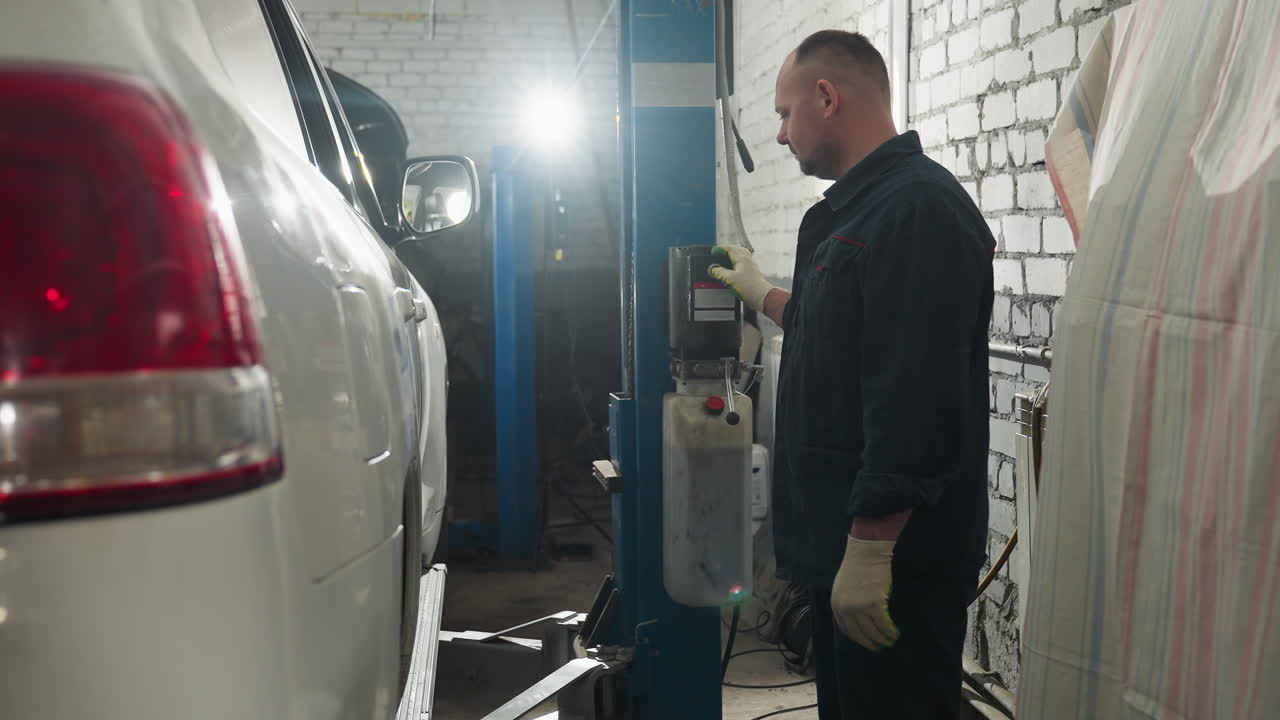 파란 유니폼을 입은 기계공은 차고에서 차를 올리기 위해 엘리베이터 컨트롤을 누르며 배경 빛은 작업 공간의 깊이와 활동을 향상시니다.