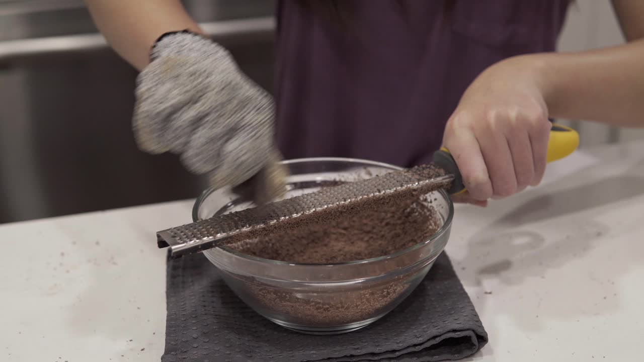 A young girl grates chocolate into a bowl for melting while making millionaire shortbread cookies while wearing safety gloves