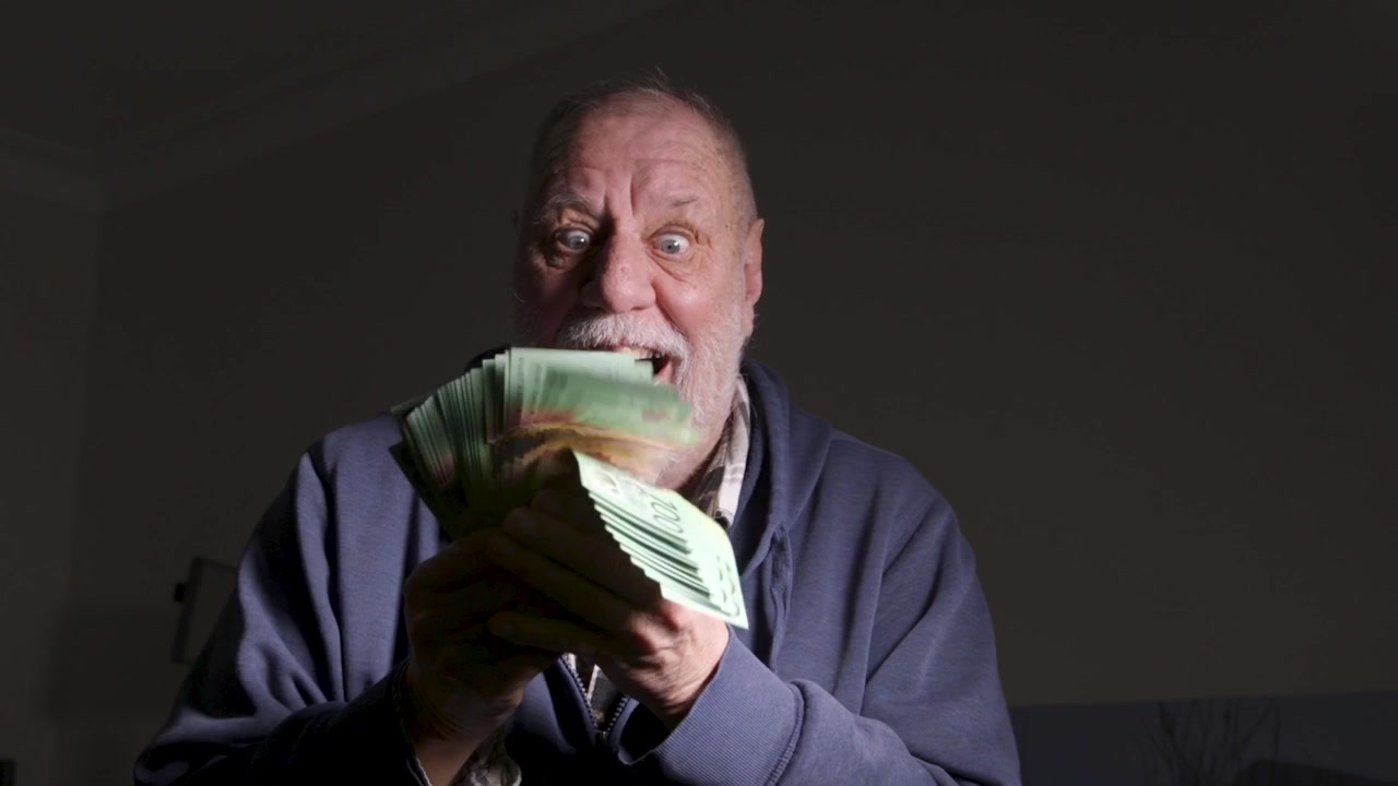 Older man in a hoodie enthusiastically fans a thick stack of green currency notes in a dark indoor room.