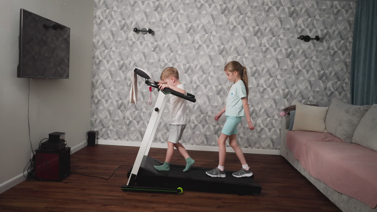 Two kids engaging in light treadmill workout inside modern living room with patterned wallpaper and wooden floor, girl walking behind boy who holds handles for balance