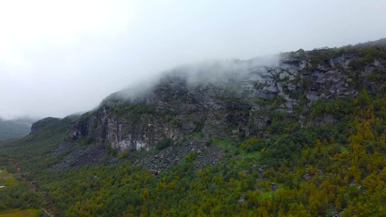 Aerial drone footage rising up and flying closer to a large mountain covered in white clouds at the top in Sweden forest nature landscape that are green during a cloudy day. Mountain is granite.