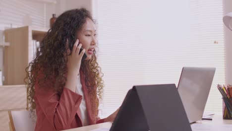 la mujer diseñadora asiática usa su teléfono inteligente para comunicarse, trabajar en proyectos y papeles, y trabajar desde casa en un escritorio con una computadora portátil.