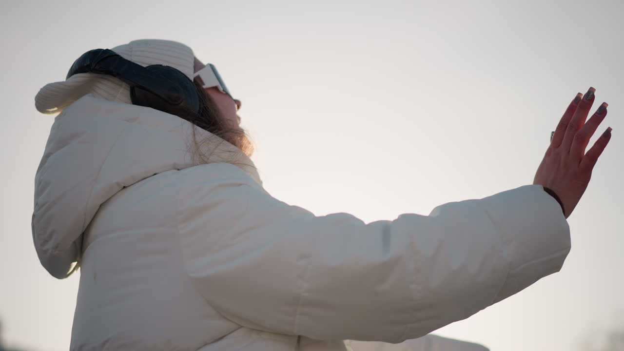 Shoulder view of creative woman smiling as she glides through park in winter outfit, wearing headphones and beanie, enjoying moment under clear sky with warm light casting gentle glow