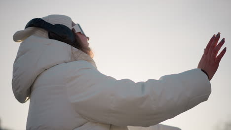 shoulder view of creative woman smiling as she glides through park in winter outfit, wearing headphones and beanie, enjoying moment under clear sky with warm light casting gentle glow