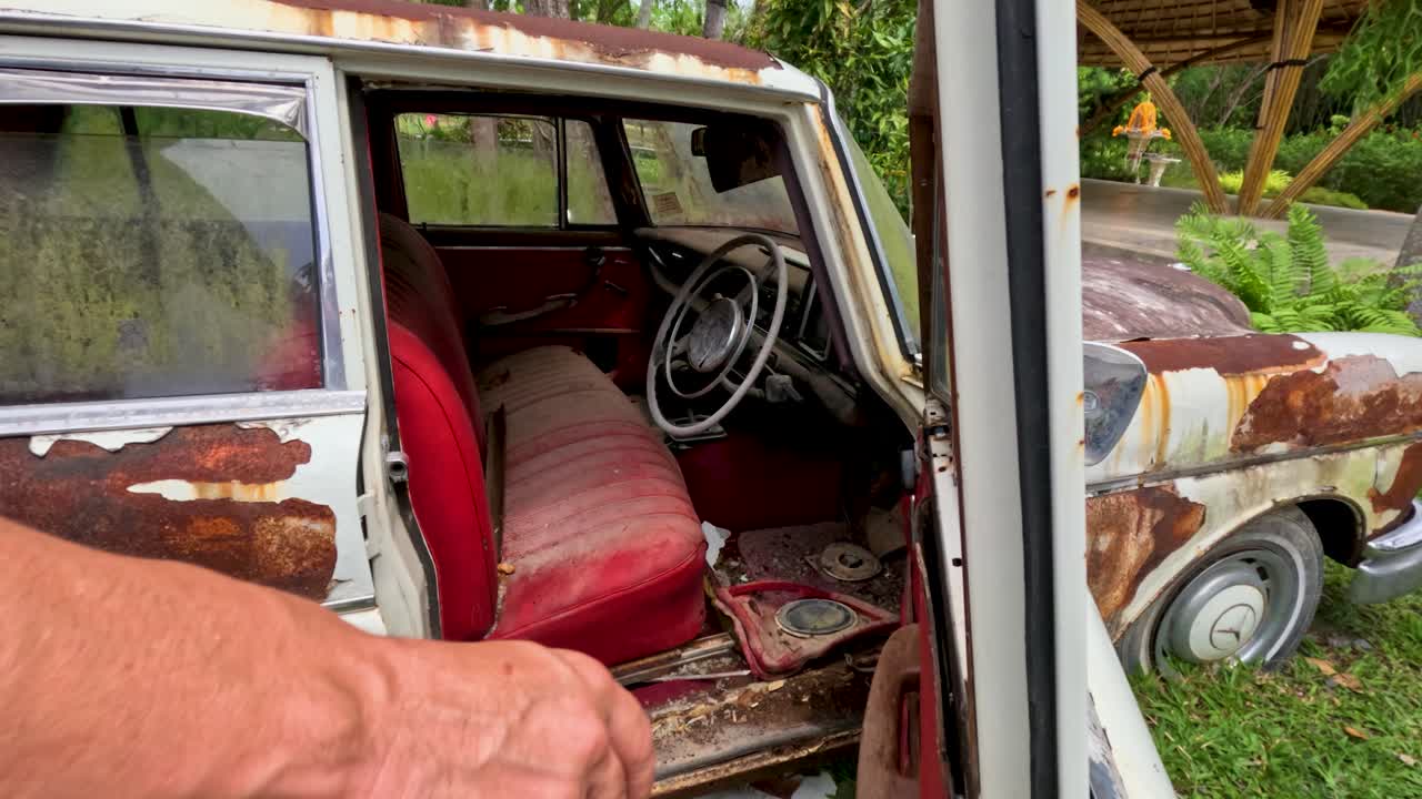 Person opens old, weathered car door revealing red interior, natural daylight, handheld camera movement