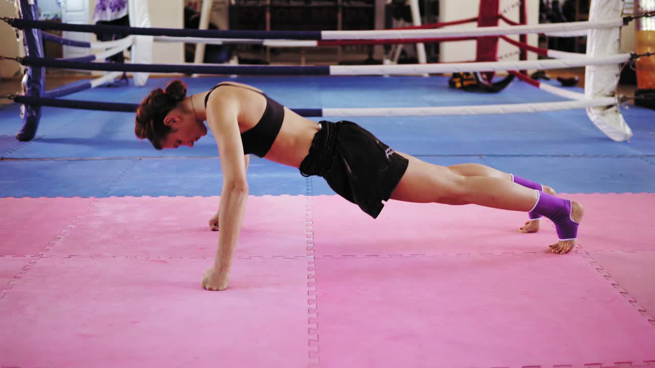 브래지어와 쇼트 입은 운동선수 여성이 무 ⁇ 과 주먹에 서서 권투 링 안의 운동 매트에서 푸시업하기 시작합니다. 힘든 힘 훈련. 4k에서 촬영