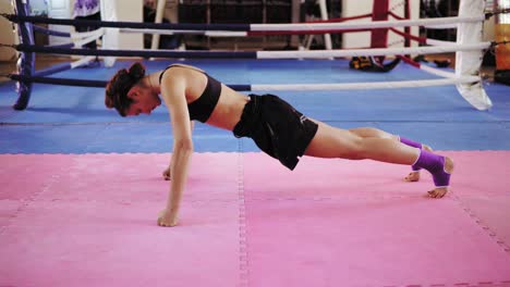 mujer atlética con sujetador y pantalones cortos de pie sobre sus rodillas y puños luego comenzando a hacer flexiones en la alfombra de ejercicio dentro del ring de boxeo. entrenamiento de fuerza duro. filmado en 4k