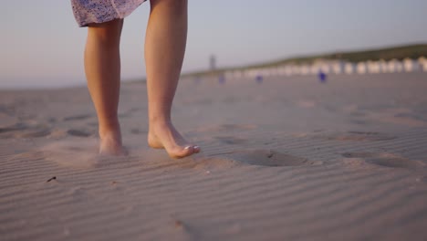 Low-close-up-dolly-shot-to-a-rising-pedestal-shot-of-a-beautiful-young-women-wearing-a-blue-summer-dress-walking-barefoot-on-a-sandy-beach-at-sunset,-slow-motion