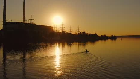 silhouette of a factory near the river. man in a boat roaming with oars at sunset. high voltage electric towers near the water against the setting sun.