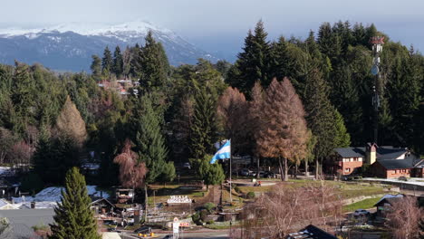 drone push-in over villa la angostura, showing the argentine flag waving at the civic monument along avenida arrayanes, framed by dense patagonian forest and snow-capped andes peaks beyond