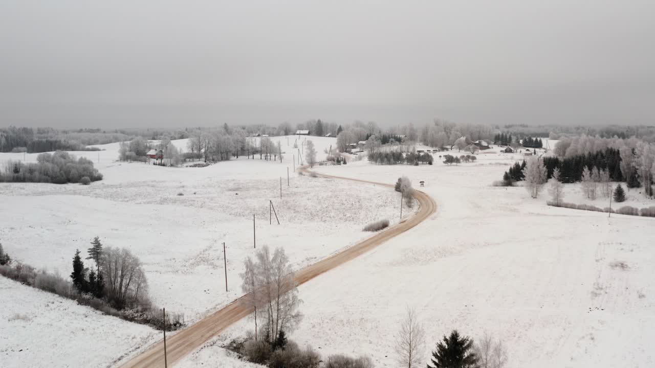 Aerial view of winding gravel road in countryside landscape with snow covered ground and frost in the trees. Winter scenery over a rural area with small community houses.