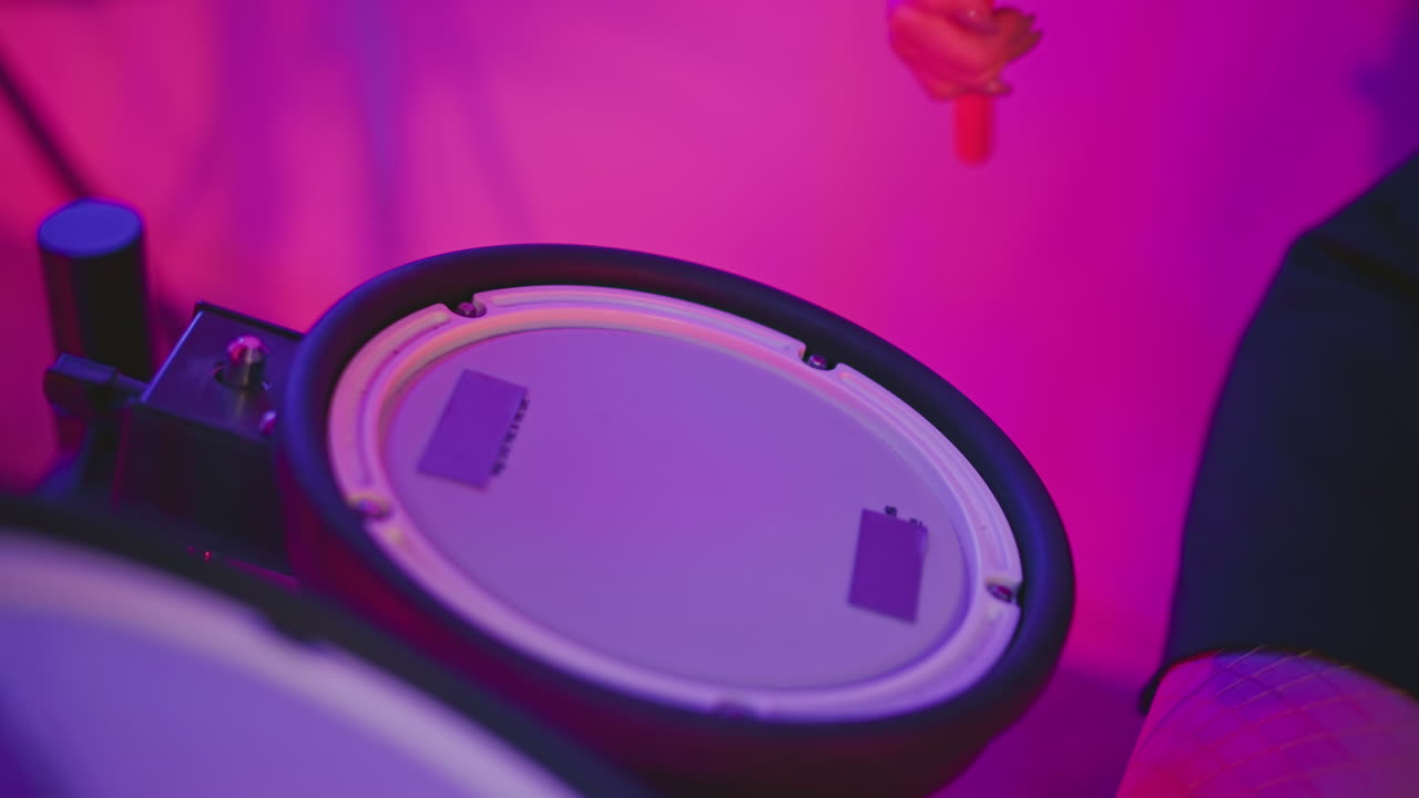 Close up of person practicing drumming skills on electronic pad in studio with glowing purple and pink lights, hand holding drumstick captured mid-motion