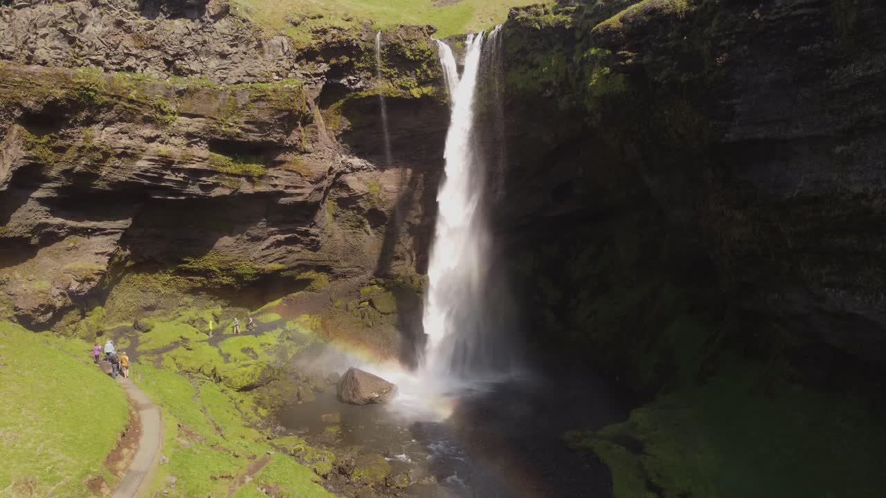 foto de un avión no tripulado con un gran ángulo retrocediendo de una hermosa cascada en islandia en un día soleado con pájaros volando y un pequeño arco iris frente a los acantilados y rocas verdes de musgo