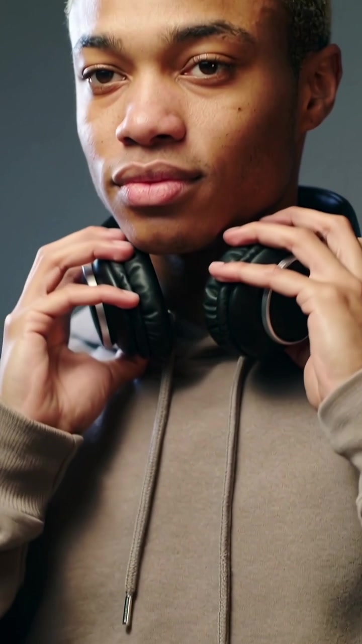 Vertical video: Lifting headphones, hooded man adjusting them at desk with controller to monitor