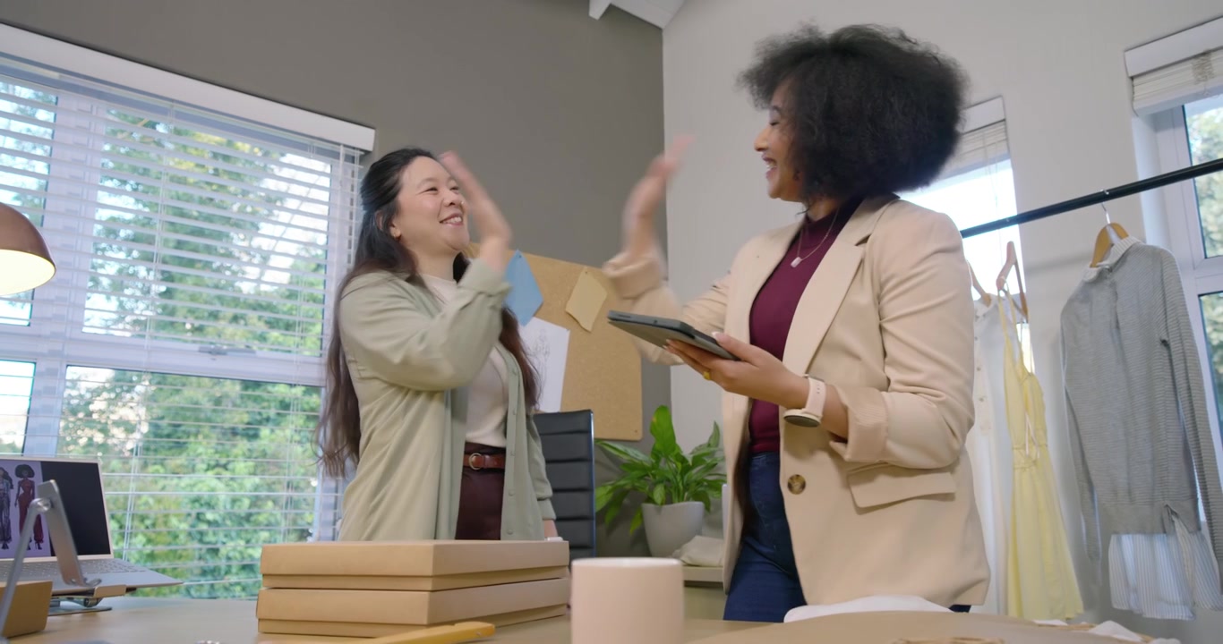 Equipe feminina diversa embalando pedidos em caixas no estúdio, embrulhando roupas após um alerta no tablet.