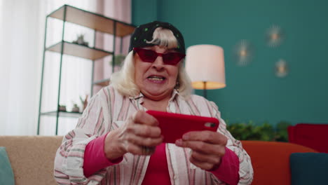 Worried-funny-grandmother-woman-enthusiastically-playing-racing-video-games-on-mobile-phone-at-home