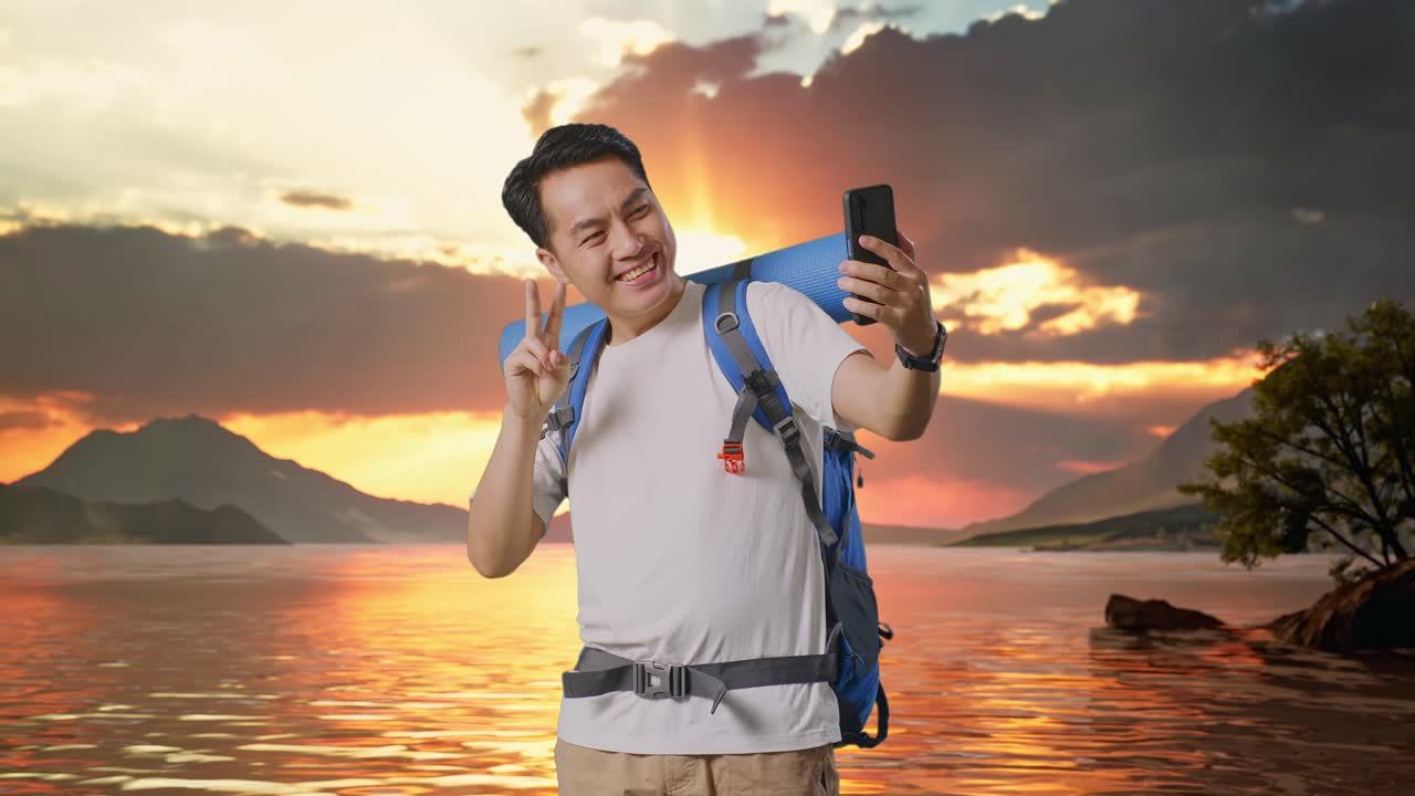 excursionista masculino asiático con mochila de montañismo mostrando paz y pulgares hacia arriba mientras toma una selfie en un lago