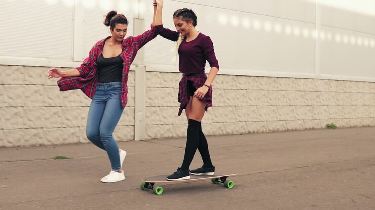 Laughing young hipster girl being taught skateboarding by a friend who is supporting her holding her hand. Slow Motion shot