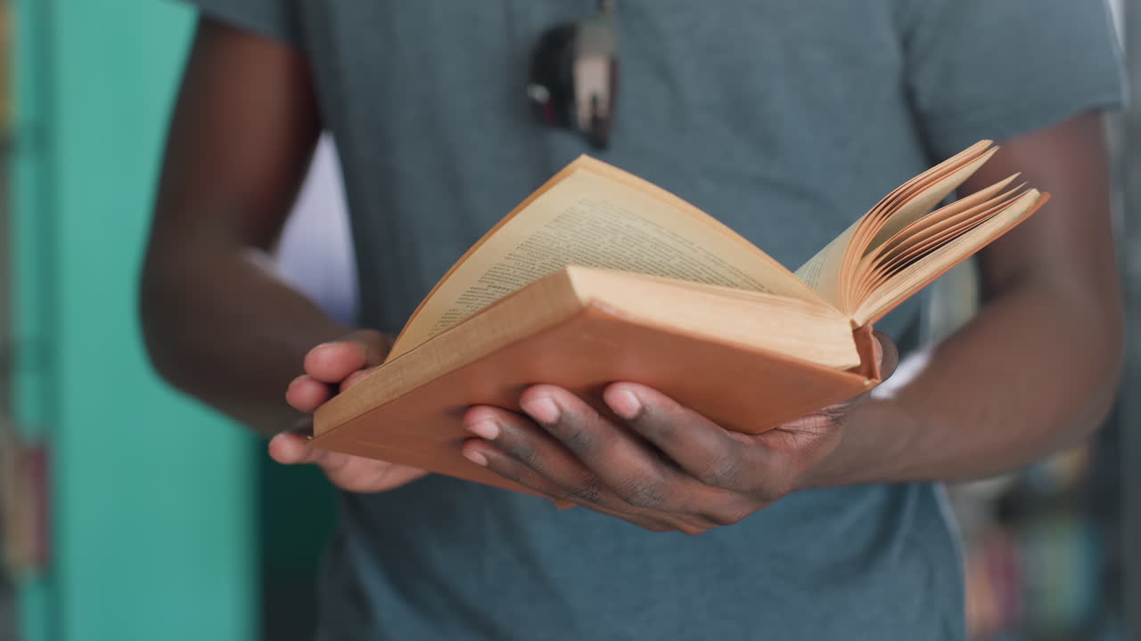 Close up student flips through vintage brown hardcover in quiet library, hands holding open pages, gentle page turn motion, sunglasses on chest, teal shelves blurred behind