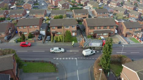 Ein-Müllwagen,-Ein-Müllsammelfahrzeug,-Fährt-Die-Straße-Hinauf,-Während-Männer-In-Einem-Mittelarbeiterviertel-Der-Stadt-Recyclingbehälter-In-Die-Müllpresse-Laden