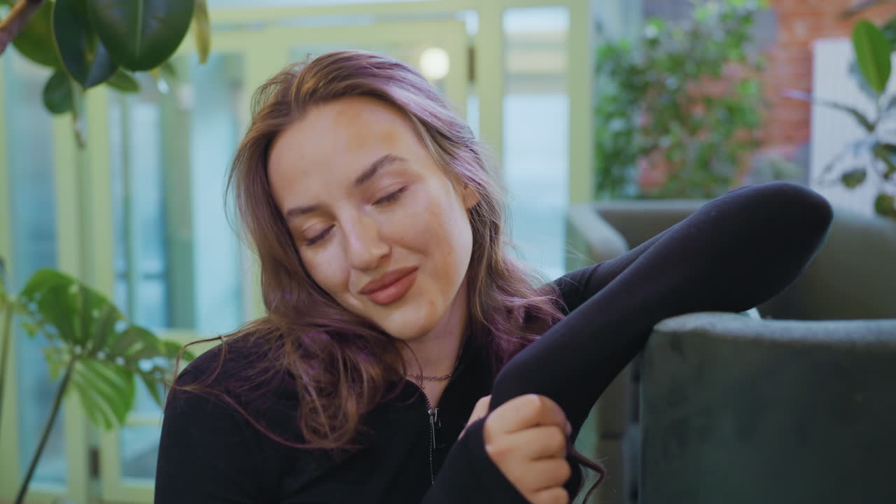Charming blond woman in black top gently leans on cushion while smiling at camera in cozy interior filled with green plants and soft daylight creating a peaceful, warm atmosphere