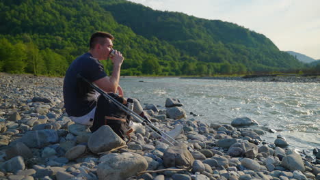 man with prosthetic leg hiking and drinking coffee by the river