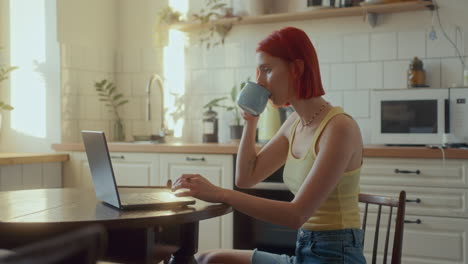 young red-haired woman sipping tea and working on laptop in sunlit kitchen