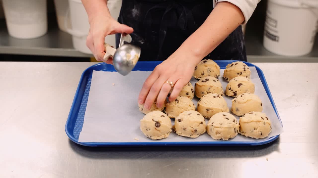 A Young Female Baker Places Cold Spherical Raw Chocolate Chip Cookie Dough Lumps on a Sheet of Wax Paper and Blue Baking Tray Inside an Industrial Professional Cooking Food Preparation Kitchen