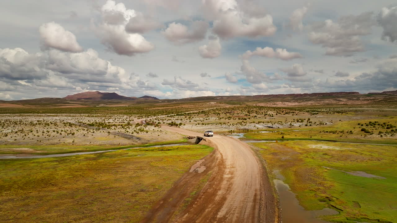 Aerial drone view of a white pickup truck driving on a dirt road in the Siloli Desert, Bolivia. The vehicle navigates the arid high-altitude landscape with mountains under a cloudy sky