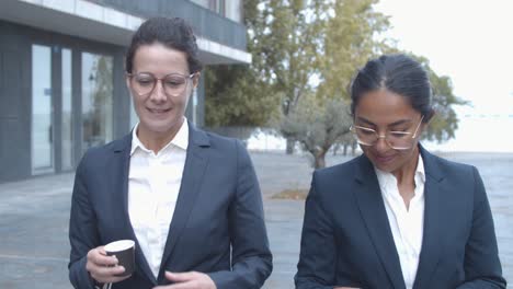 smiling female business colleagues walking at office building, drinking coffee and talking together