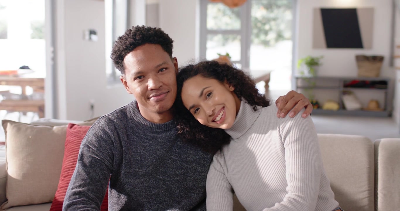 African American couple smiling at camera for portrait, nesting closer on beige sofa in living room