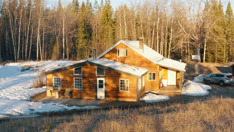 Aerial-of-quaint-wooden-cabin-house-at-sunrise-in-a-semi-snowy-forest