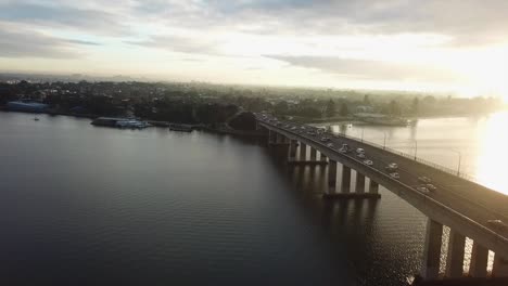 Antena:-Disparo-De-Un-Dron-Siguiendo-A-Los-Autos-Que-Cruzan-Un-Puente-Hacia-La-Ciudad-Durante-La-Hora-Pico-De-La-Mañana-Cuando-Sale-El-Sol-En-Sydney,-Australia