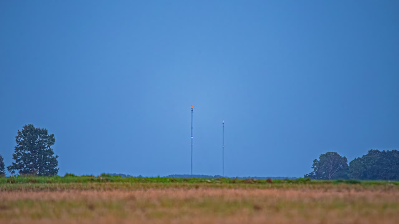 두 개의 높은 안테나가 은 하늘과 먼 나무의 배경에 서서 시골의 여름 저녁의 조용한 분위기를 포착합니다.