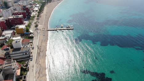 Drone-view-in-Albania-flying-over-a-beach-with-crystal-clear-blue-water-ocean,-buildings-on-the-harbor-and-green-island-on-a-sunny-day