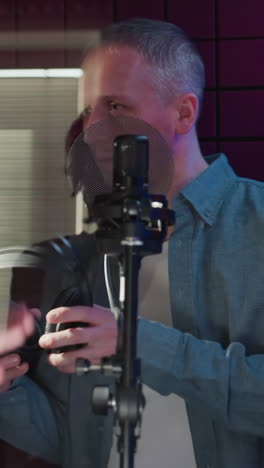 en un estudio a prueba de sonido rojo, un hombre con una camisa azul de manga larga recibe auriculares de otro hombre de negro. la interacción captura un momento de preparación para una sesión de grabación