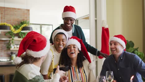 Happy-group-of-diverse-friends-in-santa-hats-celebrating-meal,-taking-selfie-at-christmas-time