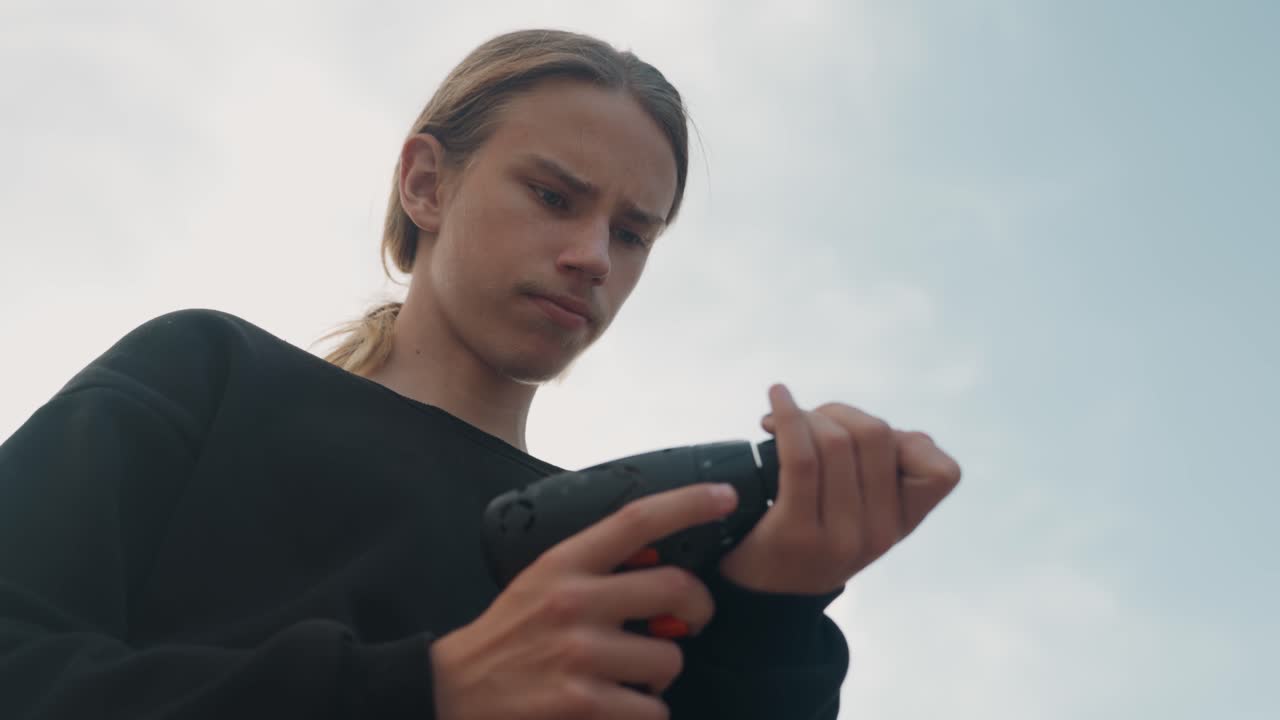 Adolescente bianco ispeziona un trapano senza fili all'aperto, apprendista falegname controlla batteria e mandrino in un'inquadratura cinematografica dal basso, sfondo di cielo tenue, espressione concentrata, mani che regolano la punta, preparazione attenta.
