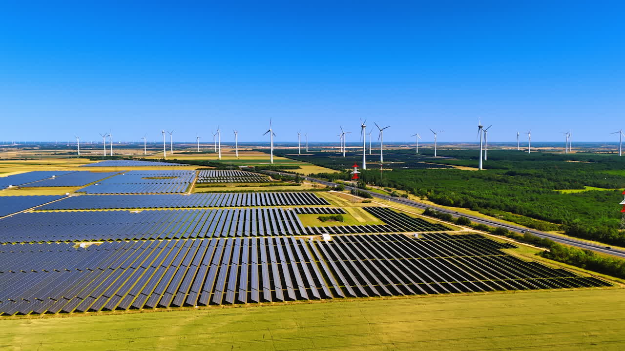 Rows of modern solar panels in the vast plantations. Wind farms at backdrop rotate slowly. Aerial view