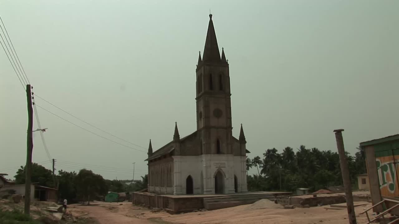 Campo lungo di una chiesa storica in stile gotico con un alto campanile e un ingresso ad arco a Elmina, in Ghana, sullo sfondo di un cielo nuvoloso con un terreno sabbioso in primo piano